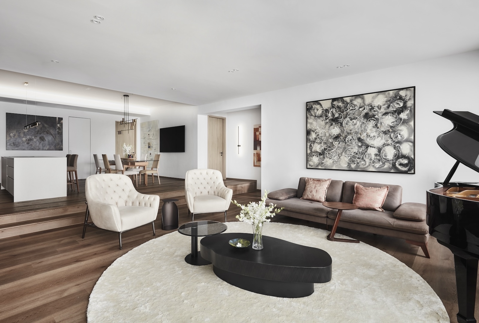 Modern living room featuring a grand piano, a round rug, a brown sofa with cushions, two beige chairs, and abstract artwork. A dining area and kitchen are visible in the background.